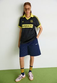 Μαύρη φανέλα Adidas με κίτρινο γιακά και ρίγες, σε συνδυασμό με μπλε σαλοπέτα με ρίγες, ροζ αθλητικά παπούτσια και μπλε κάλτσες με κίτρινες ρίγες.