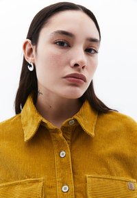 Hořčičně žlutá manšestrová košile s knoflíkovým zapínáním, širším límcem, přední kapsou a malou nášivkou s logem. Model má na sobě stříbrné kroužkové náušnice.