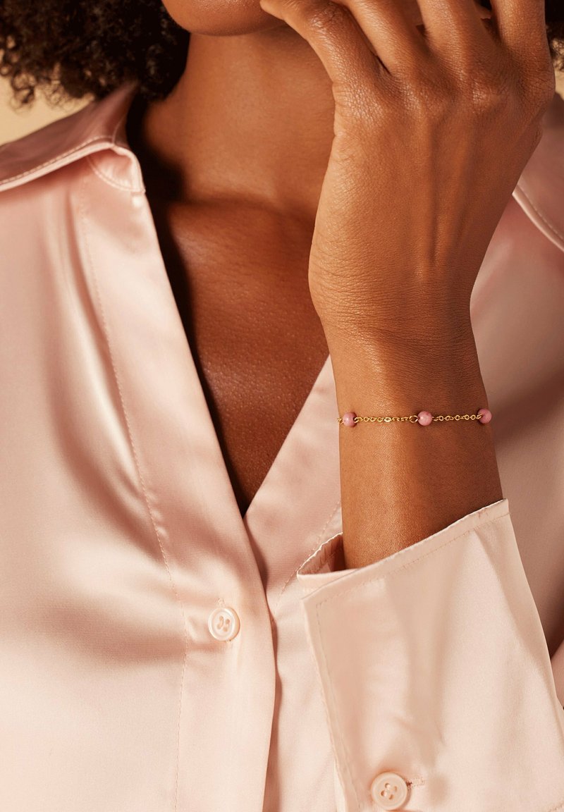 Gouden armband met paarse kralen om een pols, gecombineerd met een lichtroze satijnen shirt met een geknopt kraag- en manchetdetails.