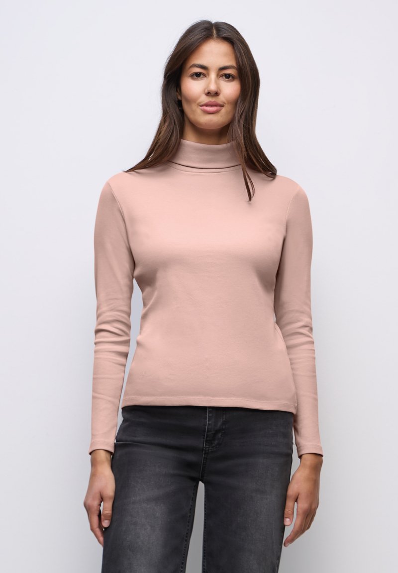 Vrouw met lang donker haar, gekleed in een lichtroze coltrui en donkergrijze spijkerbroek, staand tegen een effen lichte achtergrond.