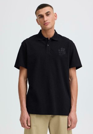 Mladi moški, ki nosi črno polo majico s kratkimi rokavi in subtilnim cvetličnim logotipom na prsih ter bež hlače, stoji na enobarvnem ozadju.