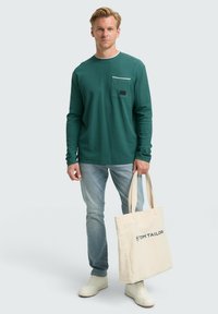 Groene blouse met lange mouwen, witte kraag, borstzak en een zwart label. Model houdt een beige schoudertas met het logo "TOM TAILOR" vast. Lichte spijkerbroek.