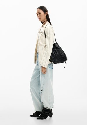 Noor naine valges triibulises bleiseris, lõdvas heledas teksapükstes, mustade kontsakingadega ja üle õla kantava musta kootud kimbukotiga.