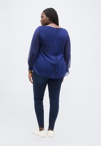 Γυναίκα με πλεξούδες, φορώντας διαφανές μπλε πουκάμισο με μακριά μανίκια, σκούρο στενό τζιν και λευκά αθλητικά παπούτσια, όρθια με την πλάτη στραμμένη σε λευκό φόντο.