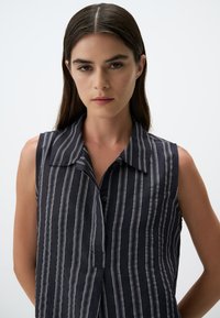 Blusa senza maniche di colore blu navy con righe verticali bianche, colletto classico e patta con bottoni; realizzata in tessuto leggero con una consistenza morbida.
