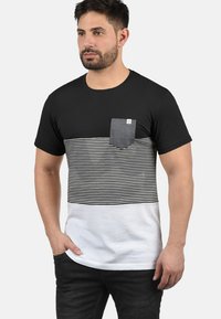 Camiseta a rayas en negro, blanco y con un bolsillo en el pecho. Con mangas cortas, cuello redondo y un diseño en bloques de color con rayas horizontales.