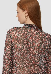 Blouse florale transparente avec un col court, présentant une base noire ornée de petites fleurs rouges et blanches, coupe ajustée et manches longues.