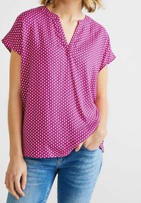 Kortärmad V-ringad topp i en ljusrosa tyg med ett geometriskt vitt mönster. Bärs tillsammans med blå jeans som har en avslappnad passform och mjuk textur.