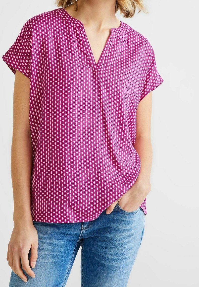 Kortärmad V-ringad topp i en ljusrosa tyg med ett geometriskt vitt mönster. Bärs tillsammans med blå jeans som har en avslappnad passform och mjuk textur.