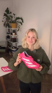 Pantof sport roz aprins cu dungi albe, partea superioară texturată și talpă amortizată. Persoana ține pantoful, prezentându-l clar.