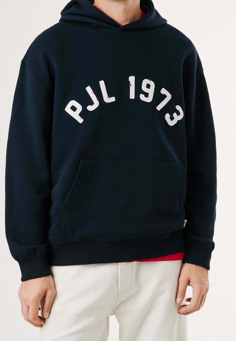 Personne portant un sweat à capuche noir avec "PJL 1973" en grandes lettres blanches et un pantalon blanc, visible des épaules aux hanches.