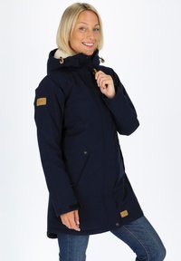 Granatowa kurtka ocieplana z kapturem wykończonym futrem, zapinana na zamek, z dwiema bocznymi kieszeniami oraz brązową skórzaną naszywką z logiem na rękawie.