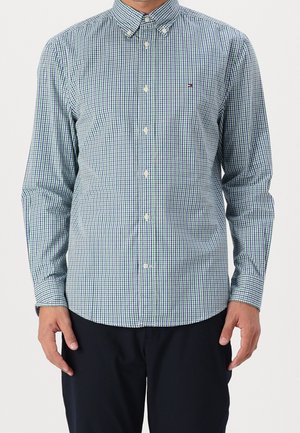 Mężczyzna w zielono-niebieskiej, kraciastej koszuli z długim rękawem, zapiętej na guziki z kołnierzykiem na guziki oraz w ciemnych spodniach.