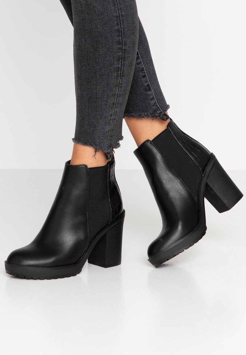 Bottines en cuir noir à talons compensés portées avec un jean skinny noir à ourlet effiloché sur fond clair.