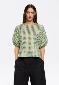 Korte mouwen top in een groen-gouden ruitpatroon, met een losse pasvorm en pofmouwen. Gecombineerd met zwarte high-waisted broek.