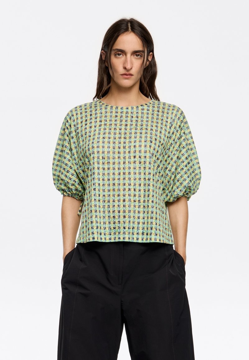 Korte mouwen top in een groen-gouden ruitpatroon, met een losse pasvorm en pofmouwen. Gecombineerd met zwarte high-waisted broek.