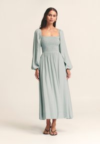 Svetlomodré šaty s dlhým rukávom, štvorcovým výstrihom a pružným sedlom. Prúdiaca látka dĺžky midi, doplnená jednoduchými sandálmi.