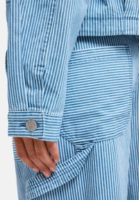 Χέρι που προσαρμόζει μπλε και λευκό πουκάμισο με κάθετες ρίγες, μακριά μανίκια με κουμπί μανσέτας και ορατή τσέπη παντελονιού με ταιριαστό ριγέ σχέδιο.