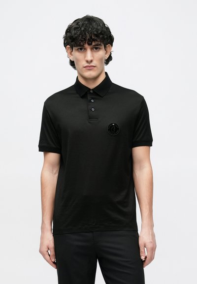 Juodas polo marškinėlis iš lygios medžiagos, turintis apykaklę, sagas priekyje ir juodą apvalų logotipo lopelį kairėje krūtinės pusėje.