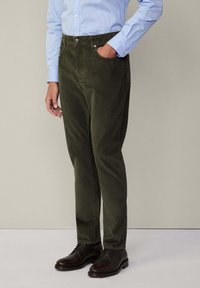 Pantaloni di velluto a coste verde oliva con una vestibilità dritta, dotati di tasche frontali e chiusura a bottone, abbinati a scarpe stringate marroni e una camicia azzurro chiaro.