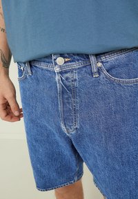 Pantalones cortos de mezclilla en un lavado azul medio, con cierre de botón y diseño de cinco bolsillos. Textura ligeramente deslavada, corte casual.