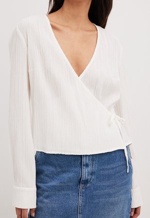 Kvinde iført en struktureret hvid slå-om-bluse med lange ærmer og blå denimjeans, bundet ved taljen på højre side.