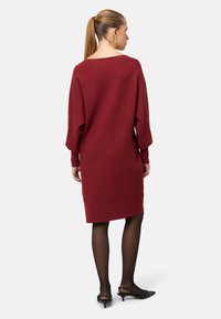 Burgunderfarbenes Strickkleid mit lockerer Passform, überschnittenen Schultern und gerippter Textur. Kombiniert mit schwarzen spitzen Absätzen und transparenten schwarzen Strumpfhosen.