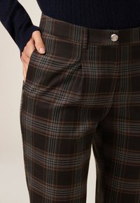 Brune ternede bukser med blå og orange detaljer. Har flad front, sidelommer og lukning med en sølvknap. Blødt, struktureret stof.