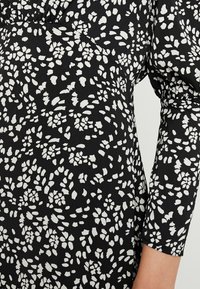 Svart klänning med ett vitt blommigt mönster, gjord av ett mjukt, texturerat tyg, med långa ärmar och samlad detalj runt halslinningen.