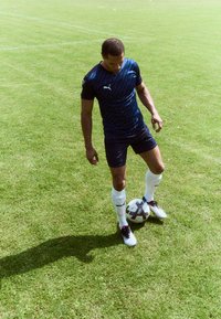 Football player in a blue striped Puma jersey and black shorts, balancing a patterned ball on the grass, wearing white socks and shoes.