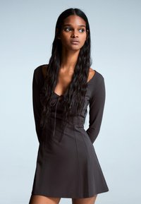 Rochie lungă maro cu mâneci lungi, cu un corset ajustat și fustă evazată. Prezintă un decolteu adânc cu detalii cu snur și material neted, elastic.