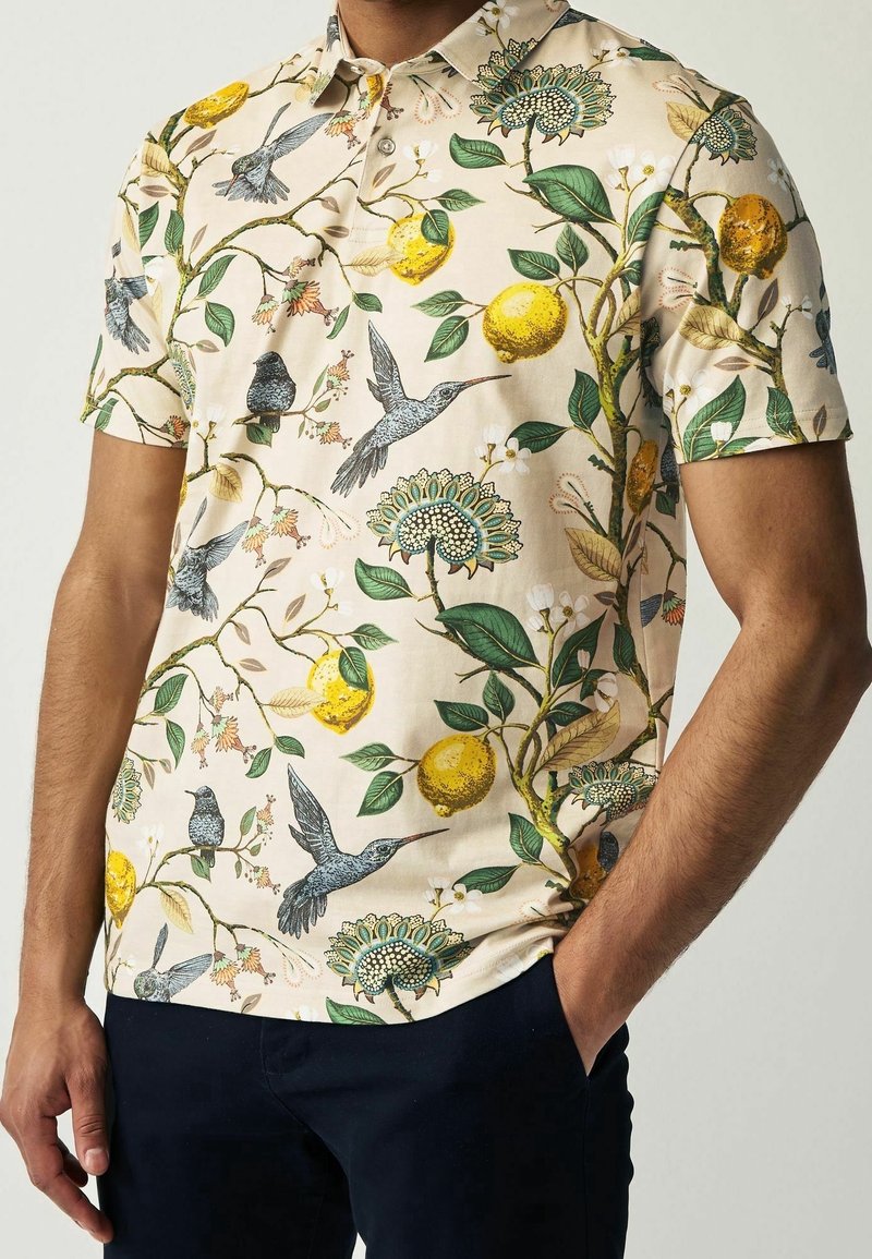 Chemise crème à manches courtes avec des feuilles vertes, des citrons jaunes et des imprimés de colibris gris portée par une personne avec la main dans la poche.