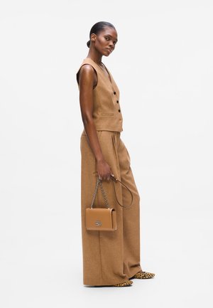 Woman standing sideways in tan sleeveless vest and wide-leg pants, holding a tan handbag with chain strap, wearing leopard print shoes.
