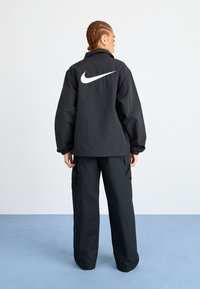 Juodas Nike švarkas su baltu logotipu nugaros pusėje. Laisvas modelis su elastingomis rankovėmis. Suderintas su atitinkamais juodais krovinių kelnėmis.