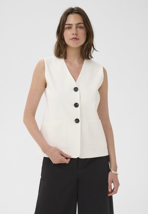 Femeie purtând un vestă albă fără mâneci, cu nasturi și buzunare frontale, și pantaloni negri, stând în fața unui fundal simplu.