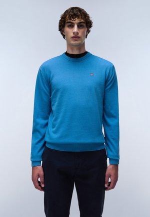 Blauwe gebreide trui met een ronde hals, lange mouwen en geribde manchetten en zoom. Voorzien van een klein logo op de borst. Gecombineerd met donkere broek.