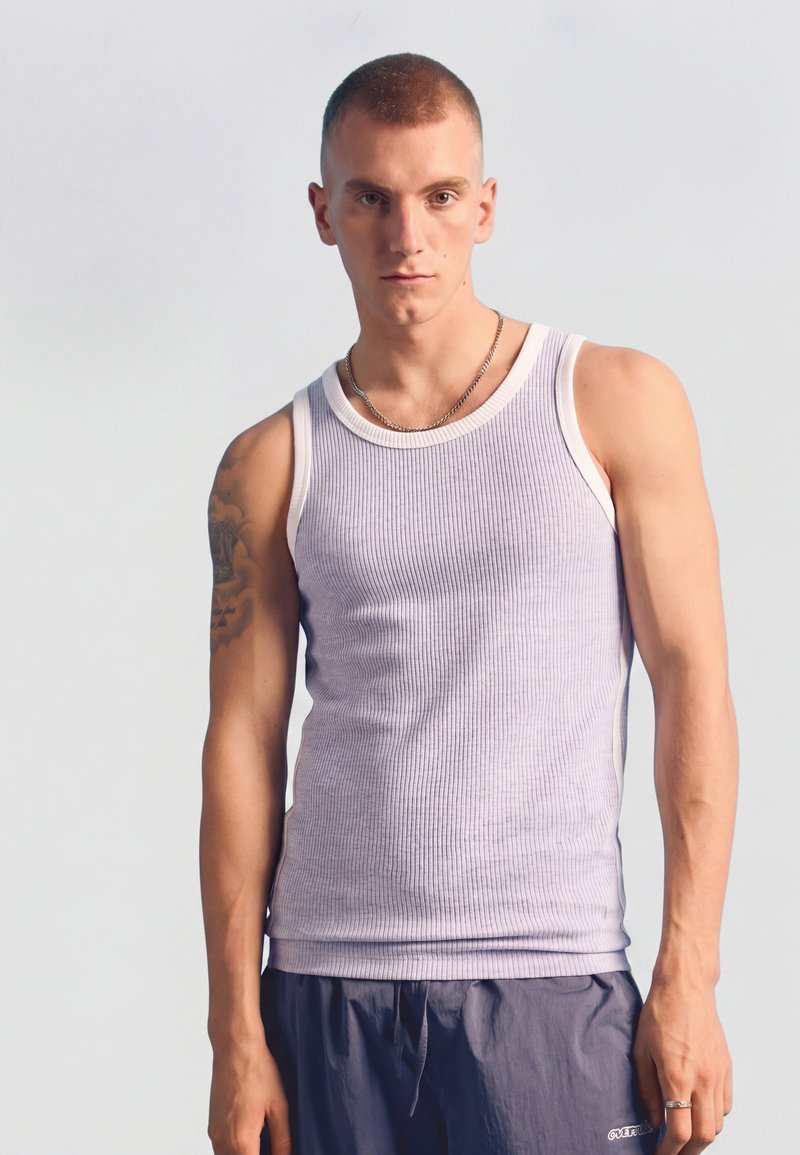 Jeune homme aux cheveux courts portant un débardeur blanc côtelé, un collier chaîne argenté et un pantalon décontracté foncé sur un fond clair uni.