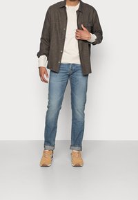 Camisa marrón de botones, sudadera crema debajo, jeans azules y zapatillas color beige. La camisa tiene un bolsillo en el pecho y una textura suave.