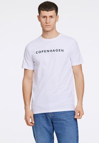 Weißes Baumwoll-T-Shirt mit kurzen Ärmeln, auf dem "KOPENHAGEN" in dunkelblauer Schrift über die Brust gedruckt ist, kombiniert mit blauen Jeans.