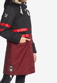 Kleurgebroken jack met een zwarte bovenkant en bordeauxrode onderkant, voorzien van rode strepen, capuchon en decoratieve hardware. Inclusief voorzakken.