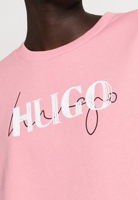 Gros plan sur une personne portant une chemise rose avec le mot "HUGO" en grandes lettres blanches au-dessus d'un texte cursif noir stylisé.