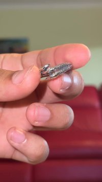 Kovový zámok s textúrovaným dizajnom, strieborným povrchom a špicatými koncami, držaný medzi prstami proti rozmazanému červenému pozadiu.