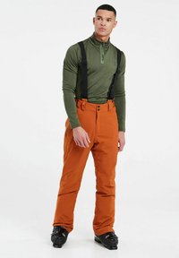 Skikleding met een groene lange mouw top en oranje skibroek met verstelbare zwarte bretels en elegante zwarte skischoenen.
