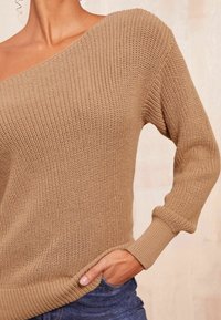 Beige gestrickter Pullover mit einem schulterfreien Design, langen Ärmeln, gerippten Säumen und einem strukturierten Muster. Eine Kombination mit dunkler Denim ist sichtbar.