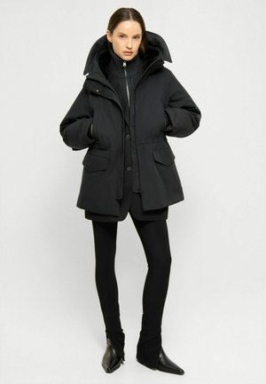 Mujer con una chaqueta acolchada con capucha negra, suéter negro con cremallera, leggings negros y botas negras con punta afilada, de pie con las manos en los bolsillos.