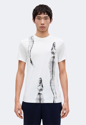 Jeune homme portant un t-shirt blanc à manches courtes avec des motifs abstraits verticaux noirs, se tenant devant un fond clair uni.