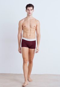 Burgunderfarbene Boxershorts mit elastischem, weißem Bund, eng anliegendem Design, mittellanger Beinform bis zum Oberschenkel und glatter Textur. Model steht barfuß.