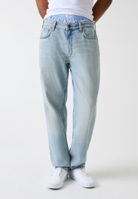 Jean en denim bleu clair avec une coupe d écontractée, doté d'un design classique à cinq poches et d'une texture subtilement délavée. Porté avec des baskets blanches.