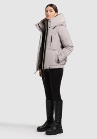 Lichtgrijze puffer jacket met een hoge kraag, voorritsen en zijzakken. Zwarte legging en kniehoge zwarte laarzen complementeren de outfit.