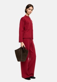 Czerwony dzianinowy strój z zamkiem z przodu, dwiema dużymi kieszeniami i elastycznym paskiem w talii. Trzyma ciemno-brązową torbę zamszową. Czarne płaskie buty.